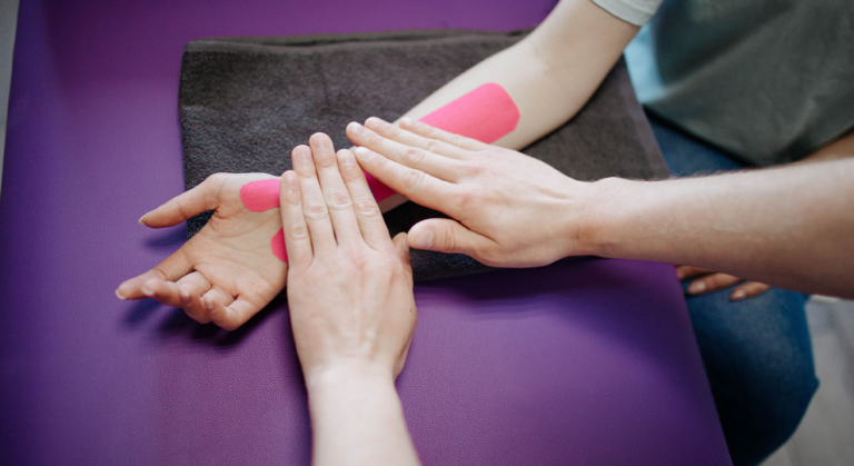 Therapist applies pink kinesiology tape to a client's forearm during a therapy session on a purple mat.