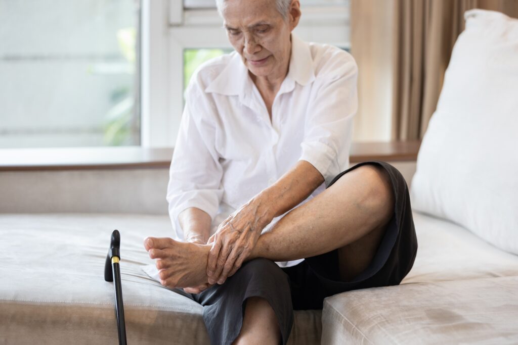 Older person sitting on a sofa, stretching their ankle with one leg raised, a cane resting on the floor beside them.