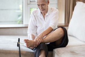 Older person sitting on a sofa, stretching their ankle with one leg raised, a cane resting on the floor beside them.